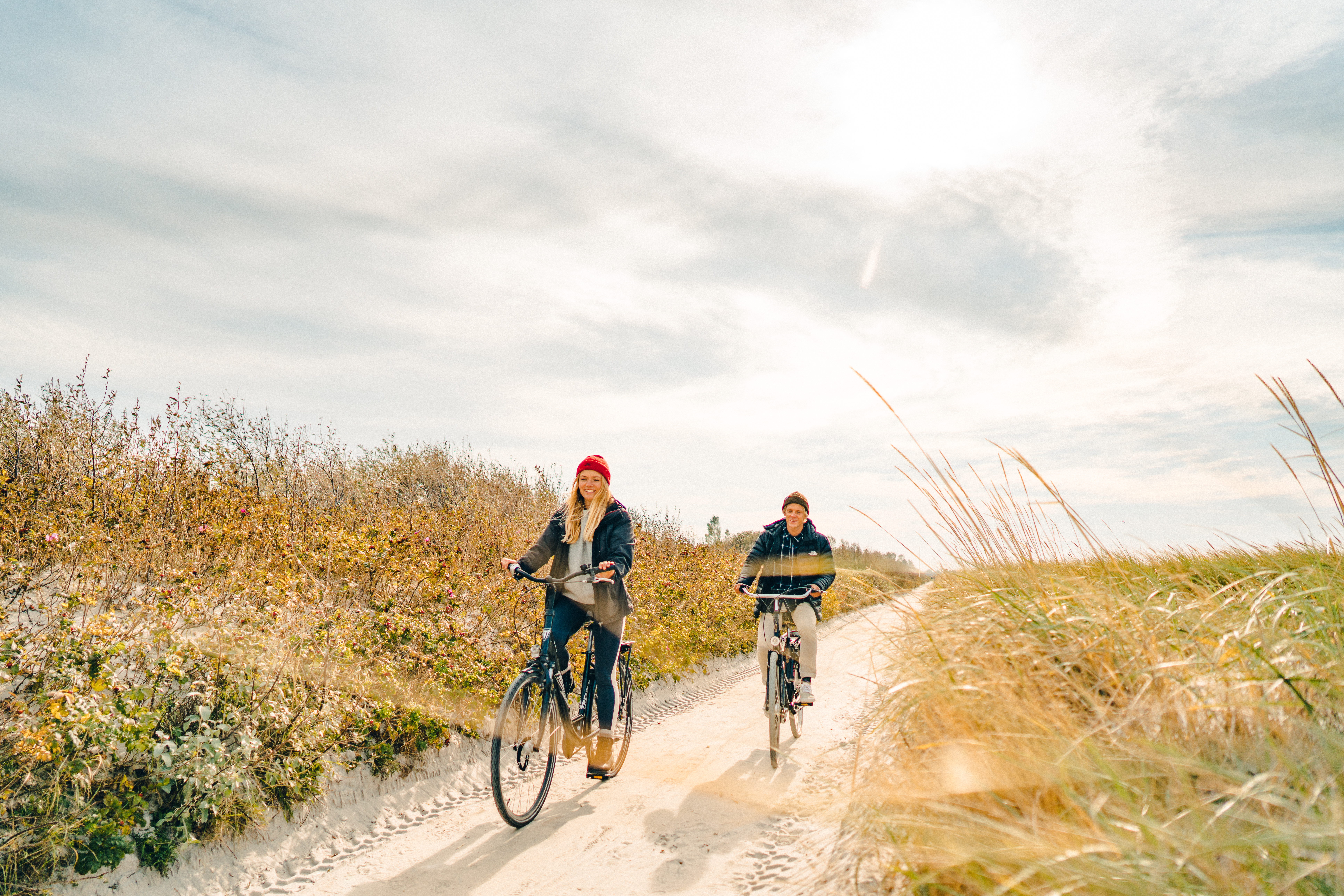 Ein Mann und eine Frau fahren auf Fahrrädern über einen Sandweg durch die Dünen. Der Weg ist umgeben von Sträuchern und Strandhafer